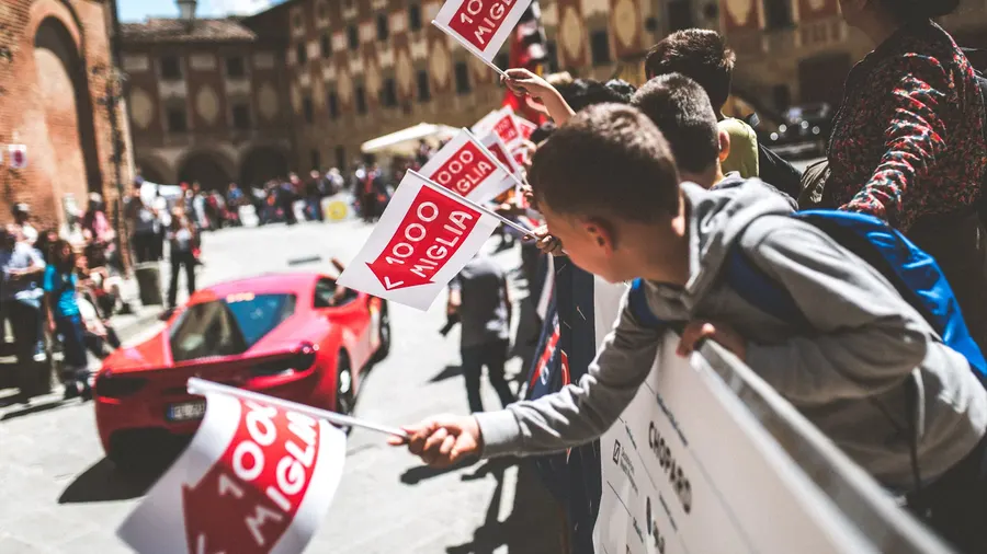 Spectateurs agitant des drapeaux Mille Miglia au passage d'une Ferrari classique dans une place de village italien